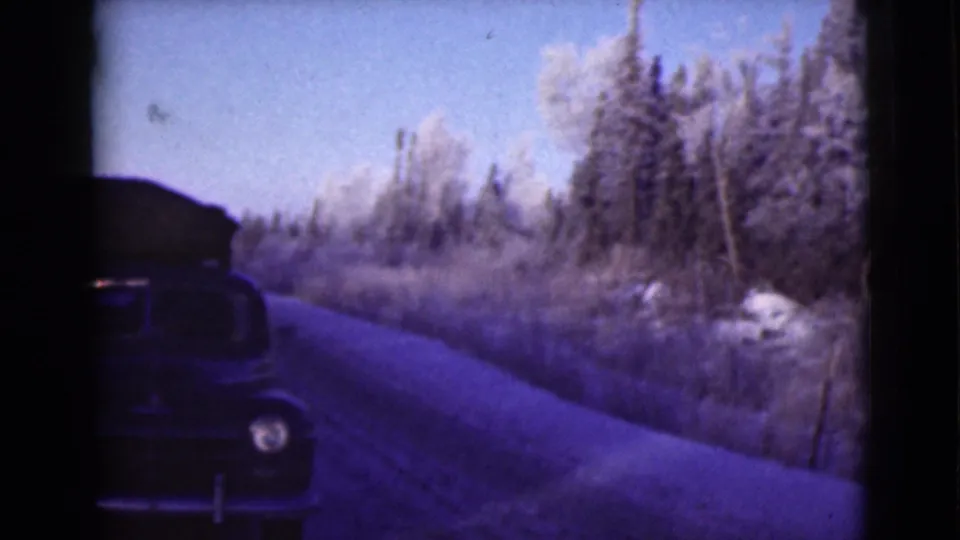 The dark car continues past the camera on a narrow Alaska road, with roof baggage silhouetted against snow-covered trees.