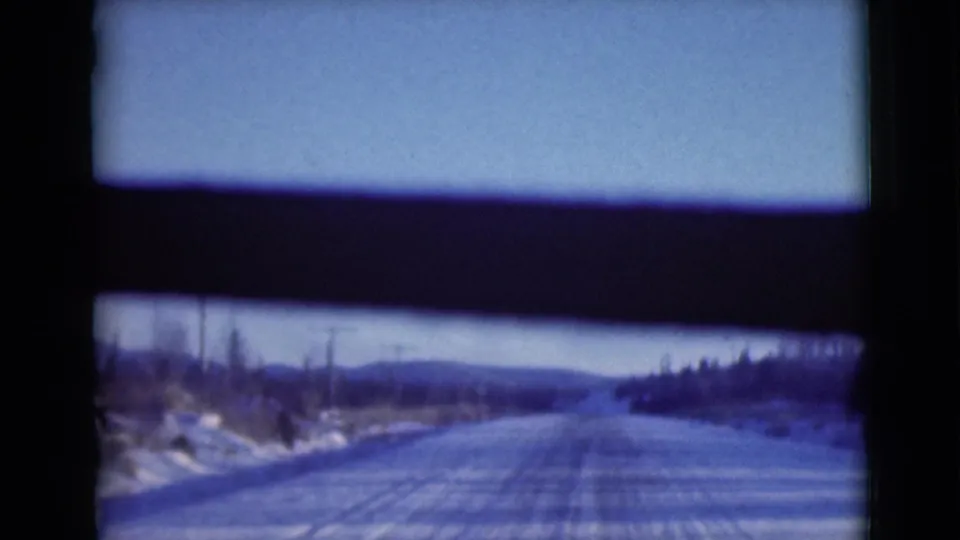 Vehicle-perspective frame of a snowy Alaska road leading toward low hills, with snowbanks, roadside trees, and dark interior framing.