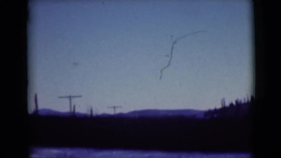 1946 Alaska road scene with utility poles, distant hills, dark tree line, and visible scratches on 8mm film.