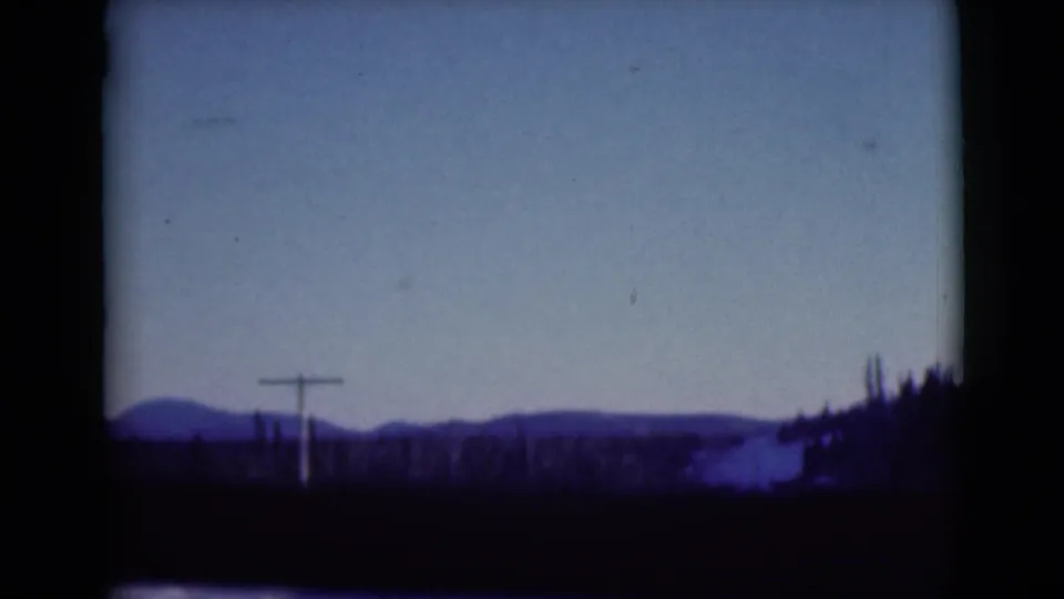 1946 Alaska 8mm frame with utility poles, distant snowy hills, dark trees, and a vehicle-window edge along the foreground.