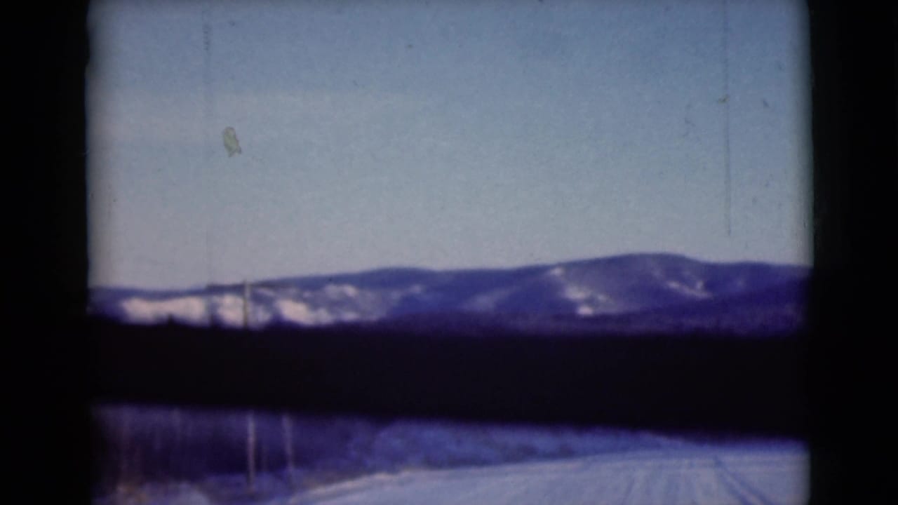 1946 Alaska home-movie frame showing a snow-covered mountain ridge through a car window, with roadside snow and a utility pole visible.