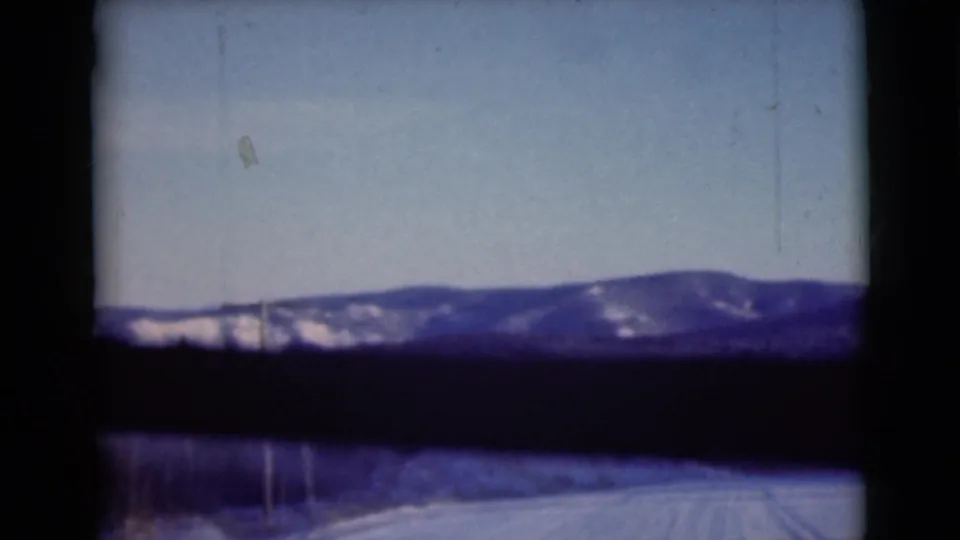 1946 Alaska home-movie frame showing a snow-covered mountain ridge through a car window, with roadside snow and a utility pole visible.