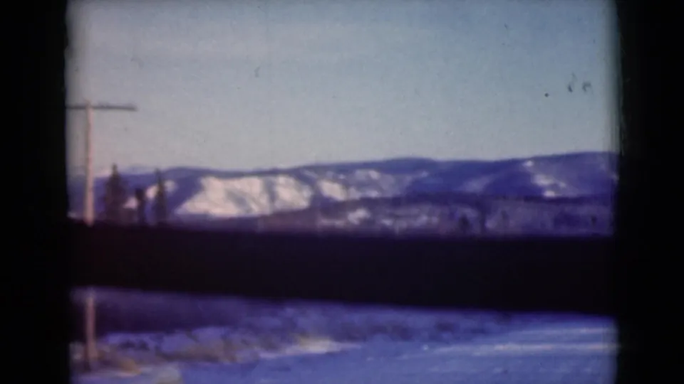 A moving-car view of Alaska winter terrain in 1946, with snow on distant hills, a dark window bar, and roadside poles.