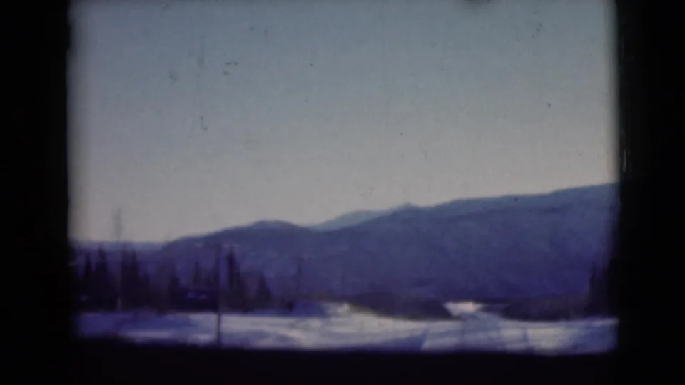 Alaska winter mountains in a 1946 8mm frame, with dark roadside trees, utility poles, snow patches, and a broad pale sky.