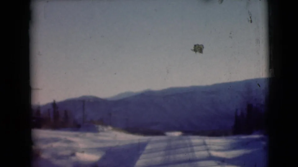 1946 Alaska home-movie frame with snowfields, a road or track, sparse trees, and distant mountains beneath a pale winter sky.