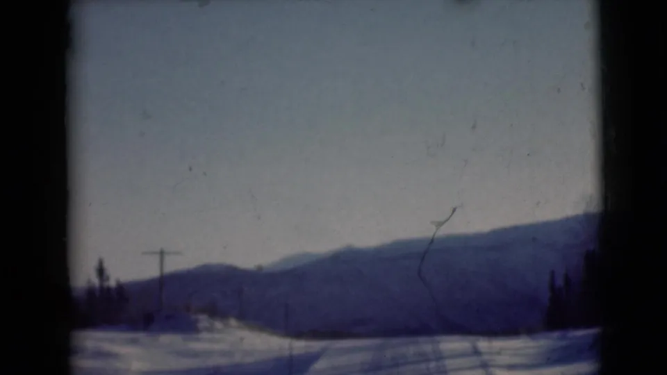 1946 Alaska 8mm frame showing snow-covered ground, a utility pole, evergreen trees, and hazy mountains beyond.