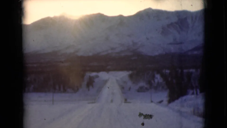 1946 Alaska 8mm frame from a moving vehicle on a snow road facing low sun, mountains, and roadside trees.