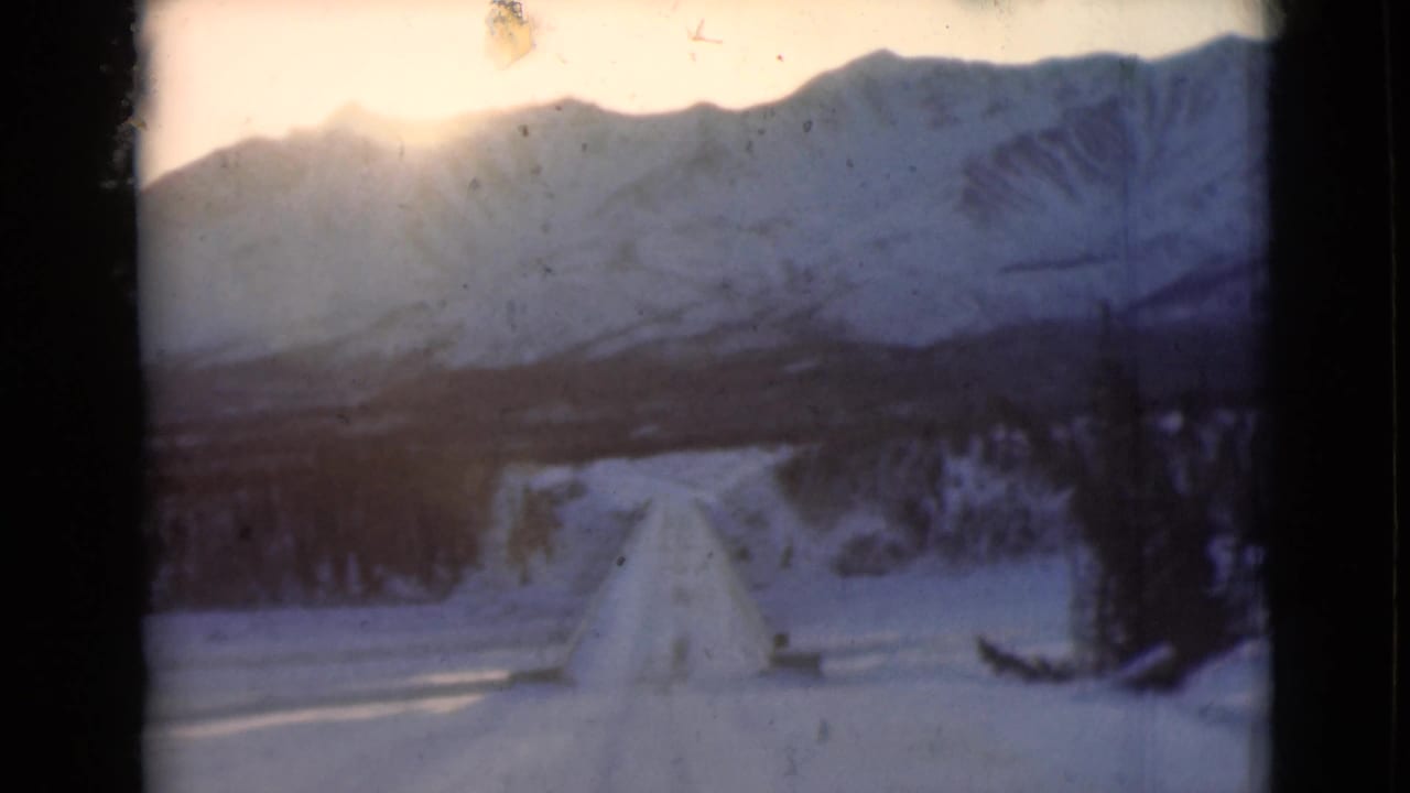 Snowy road running toward distant mountains with dark evergreen trees at the roadside.