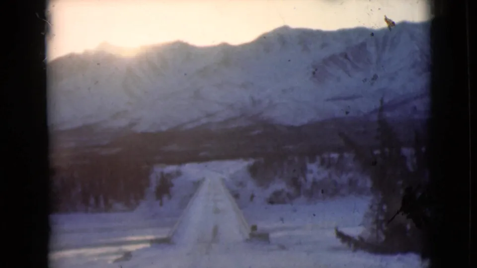 Forward-looking 8mm view of an Alaskan snowy road stretching toward mountains and roadside evergreens.