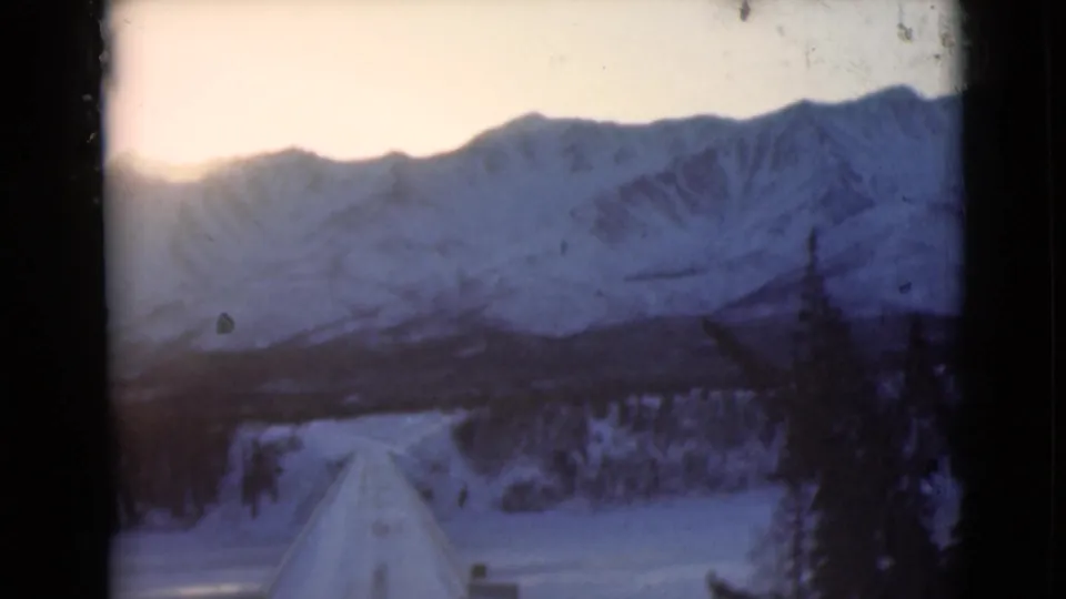 Vintage 8mm frame from Alaska showing the road rising into a snowy foreground below a broad mountain ridge.