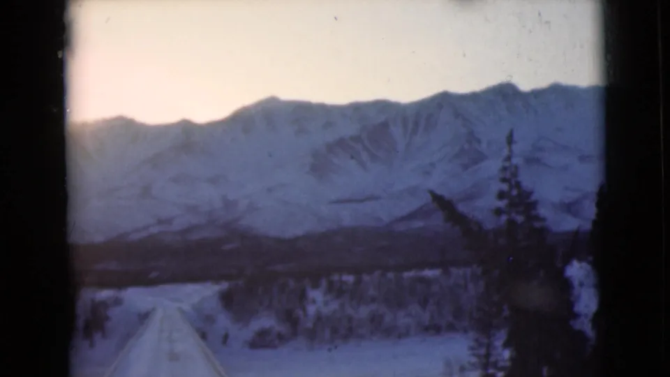 Alaska 1946 home-movie still with a snow road, evergreen tree line, and blue-toned mountains under a bright sky.