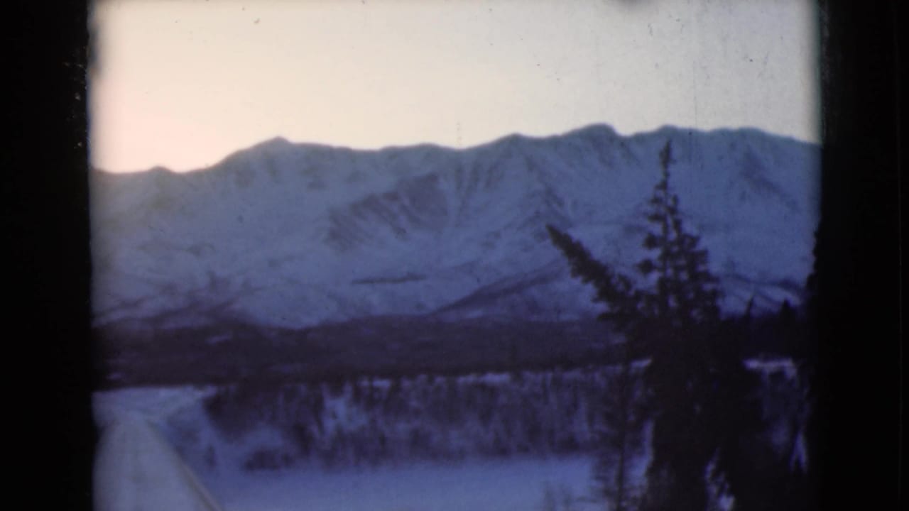 1946 8mm home-movie frame of an Alaska winter mountain range with snow-covered slopes, pale sky, and dark evergreen trees in the foreground.