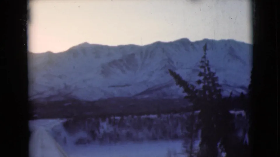 1946 8mm home-movie frame of an Alaska winter mountain range with snow-covered slopes, pale sky, and dark evergreen trees in the foreground.