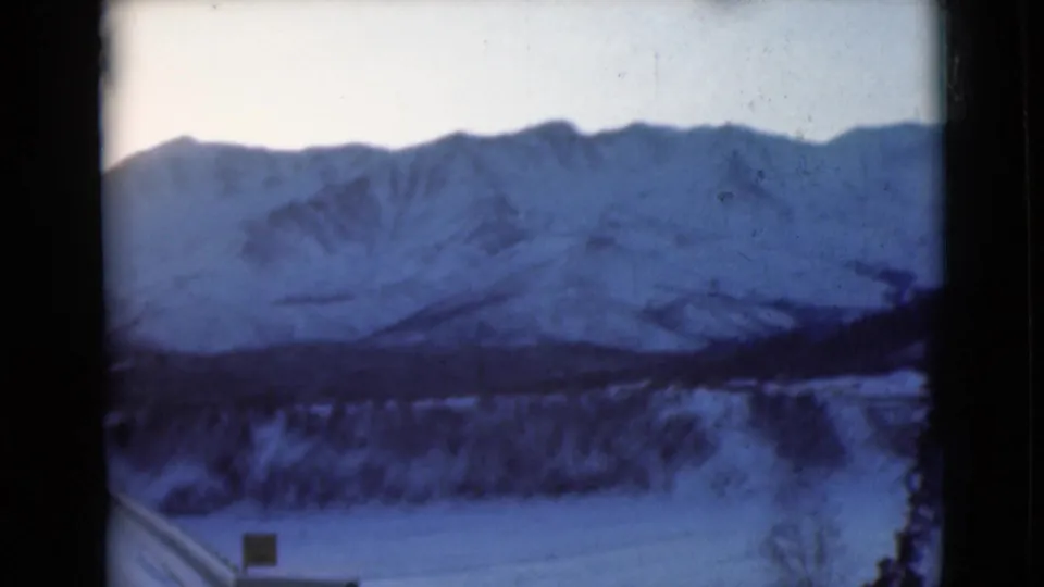 1946 8mm Alaska winter frame with a snow-covered road or track in the foreground, dark trees, and a snow-capped mountain range beyond.