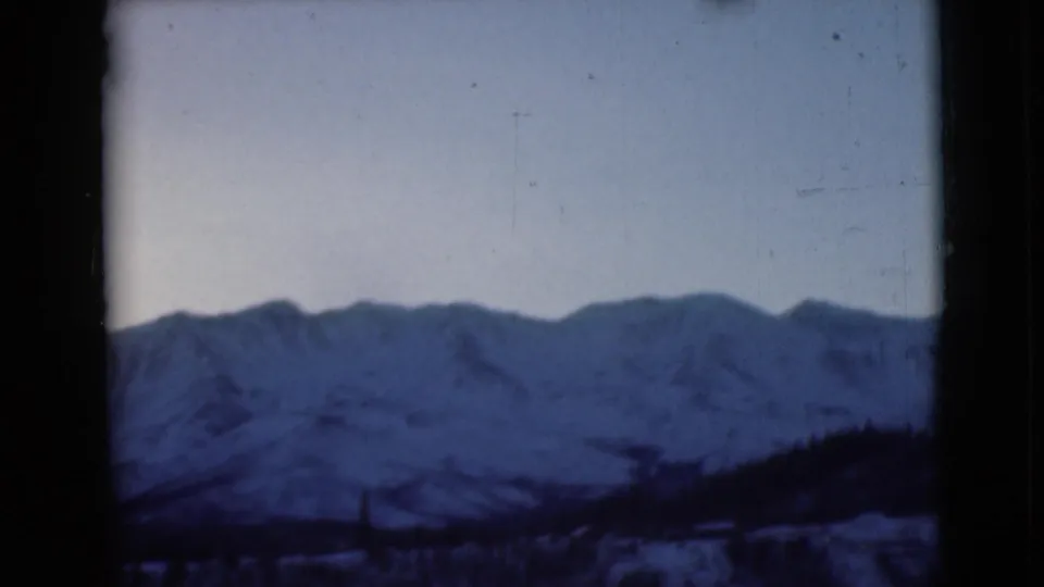 1946 Alaska home-movie frame showing a snow-covered mountain range below a pale winter sky, with a dark sloping foreground and rounded 8mm frame edges.