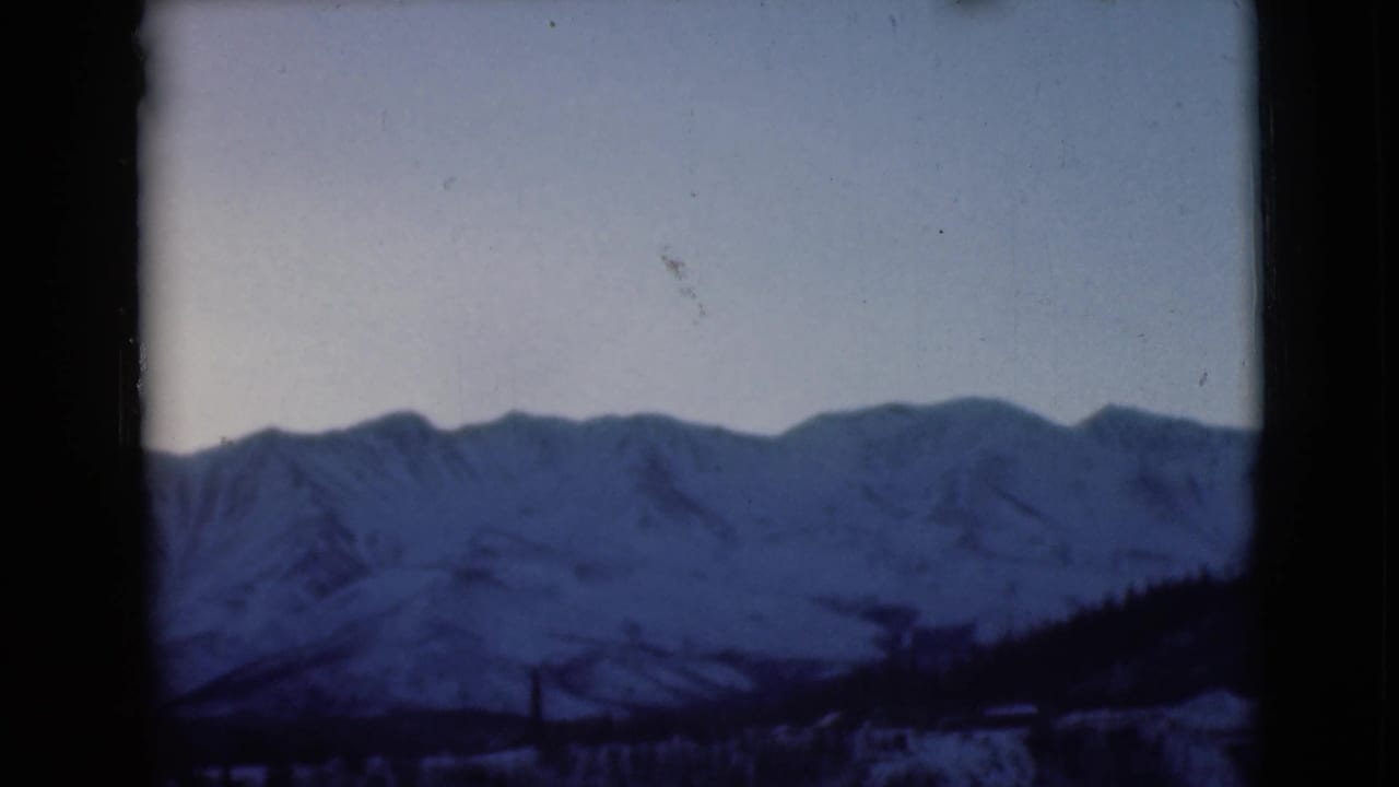 1946 Alaska 8mm frame showing distant snow-capped mountain peaks beneath a pale sky with dark borders around the view.