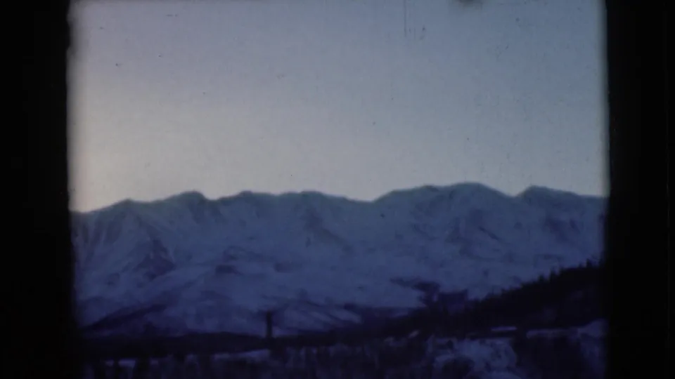 1946 Alaska home-movie still with snow-covered mountains, pale sky, dark framing, and a shadowed lower slope.