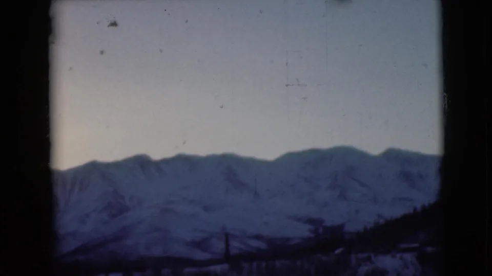 1946 Alaska 8mm frame showing distant snowy mountains through a dark window edge, with soft blue tone and film grain.