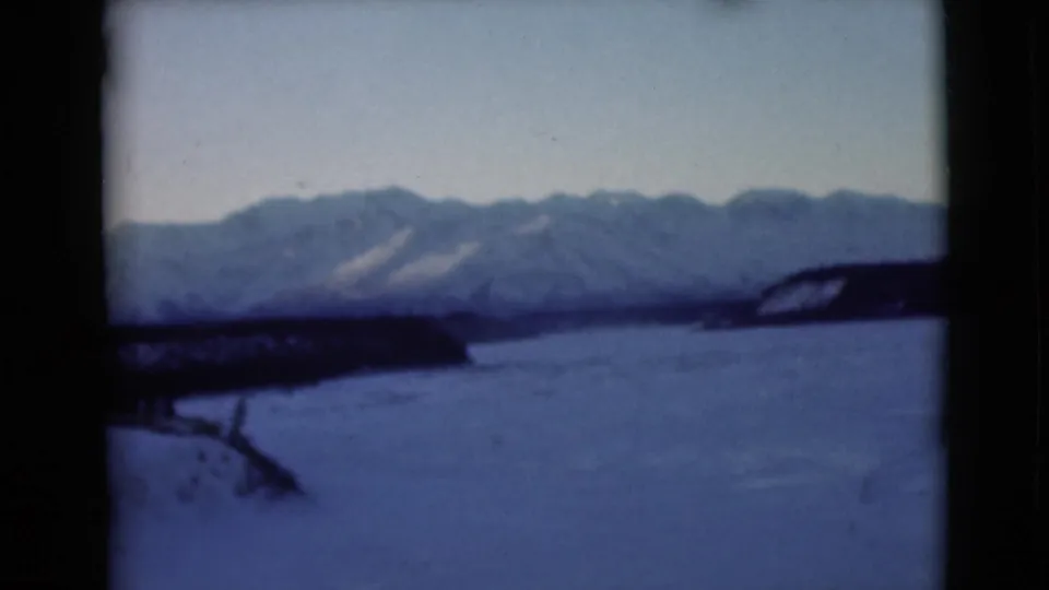 1946 Alaska 8mm frame with a frozen water surface, dark wooded shore and snow-covered mountains under a pale sky.
