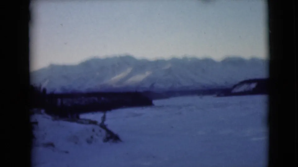 Blue-tinted 1946 Alaska 8mm view of a frozen river or lake, dark shore, sparse trees and distant snowy mountain range.