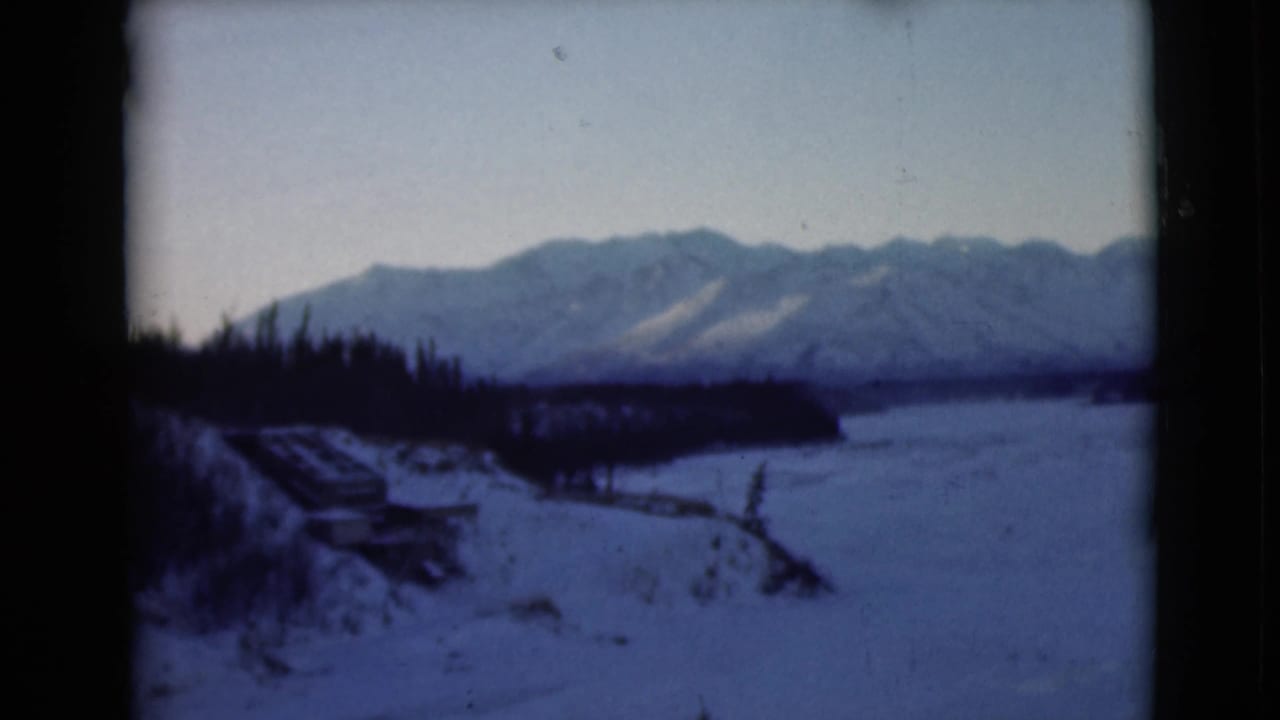 1946 Alaska 8mm frame showing a snowy foreground, dark evergreen line, and distant mountain ridge under a pale winter sky.