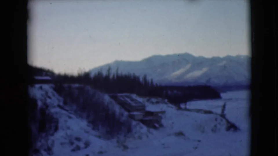 1946 Alaska 8mm frame from a moving viewpoint, showing a snowy slope, evergreens, distant mountains, and a white valley or frozen water area.