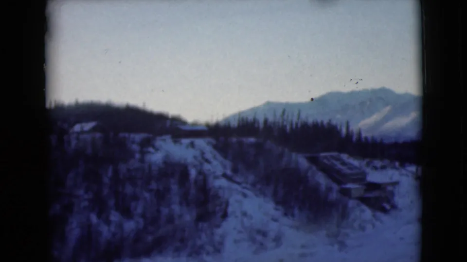 1946 Alaska 8mm frame showing a snow-covered hillside, dark trees, and a distant mountain ridge from a moving vehicle perspective.