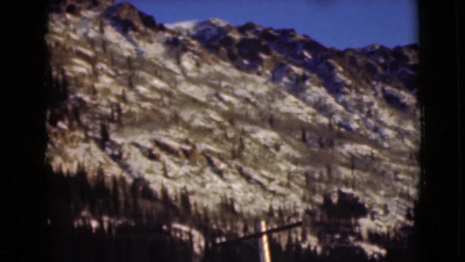1946 Alaska 8mm frame showing a sunlit rocky mountain face with snow patches, scattered evergreens, and a darker tree line at the base.