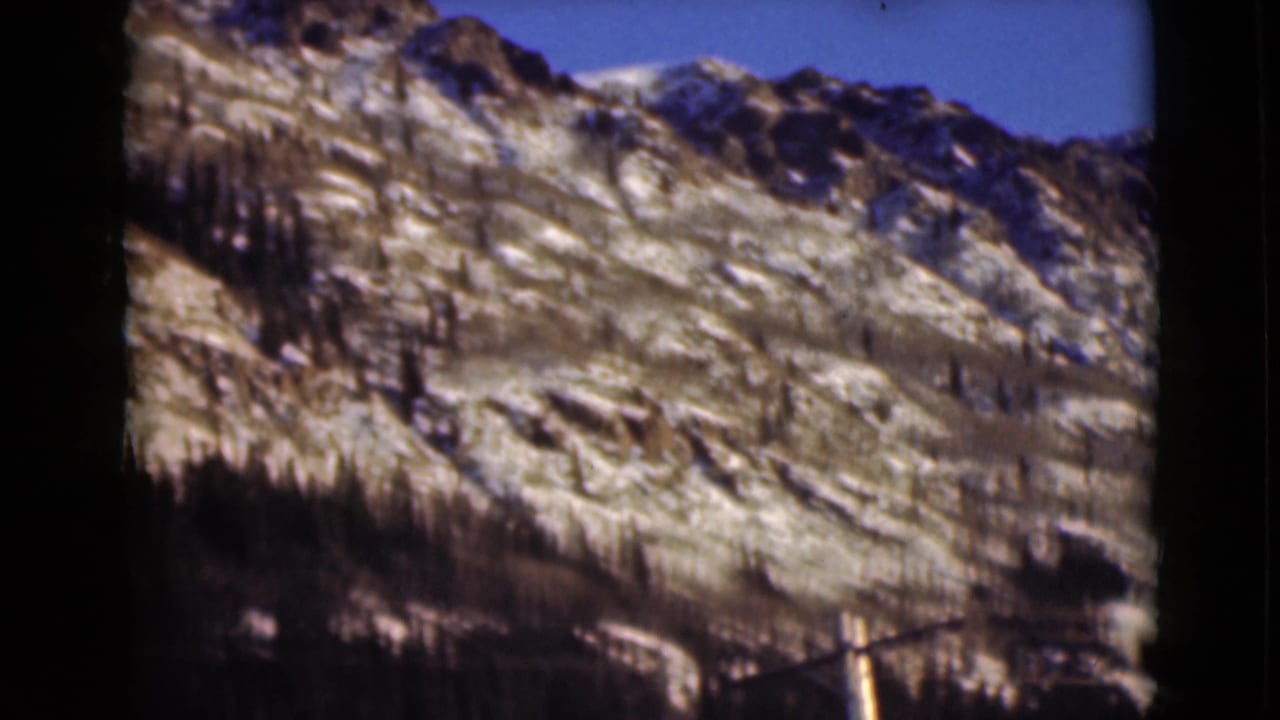 1946 Alaska 8mm frame showing a snow-dusted rocky mountain slope with scattered dark evergreens under blue sky.