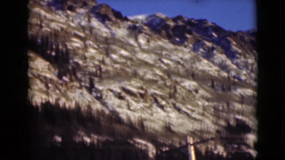 1946 Alaska 8mm frame showing a snow-dusted rocky mountain slope with scattered dark evergreens under blue sky.