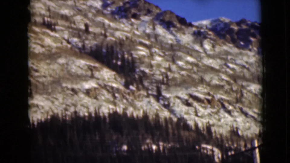 1946 Alaska 8mm view of a rugged hillside with patchy snow, dark conifer trees, and a clear blue sky.