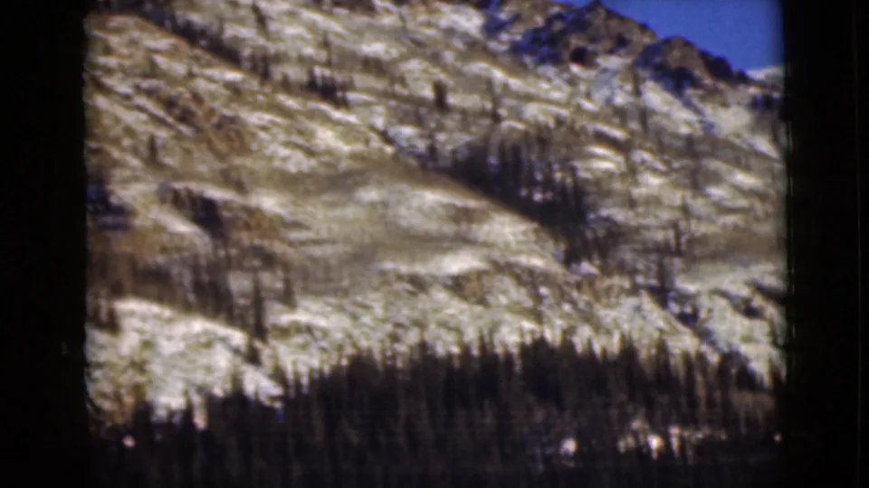 1946 Alaska home-movie frame with a pale rocky slope, scattered snow, and a dark evergreen band near the lower edge.