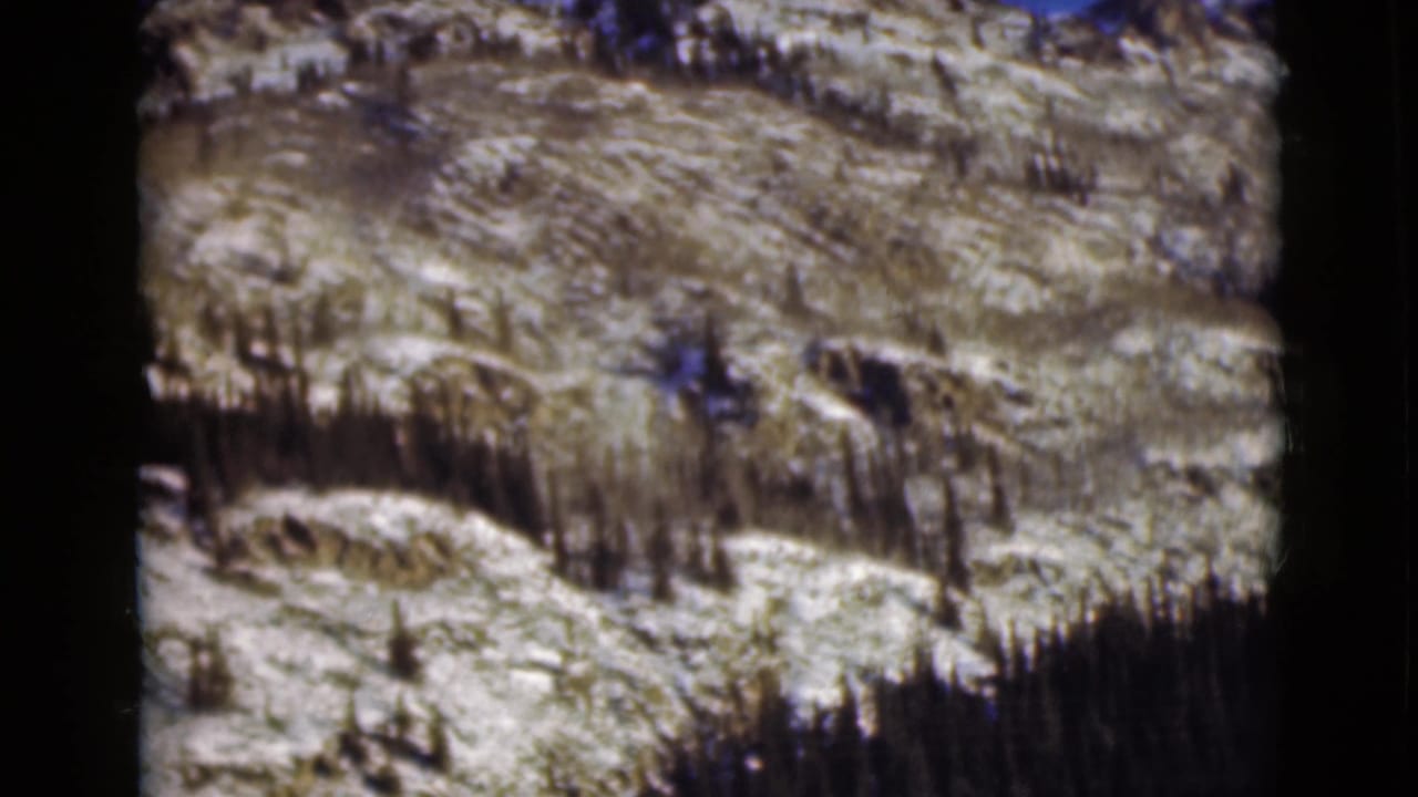Snow-dusted rocky mountainside with sparse dark trees and film gate edges.