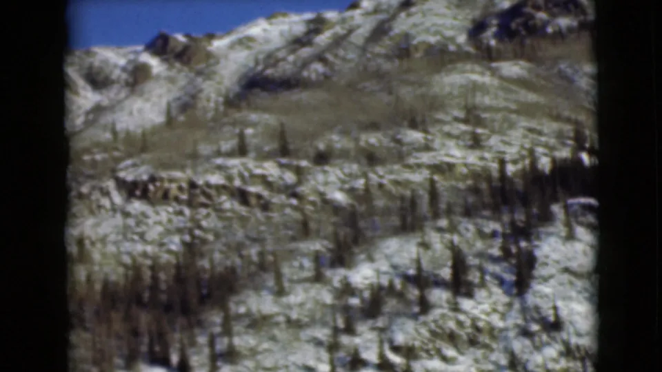 1946 Alaska frame showing a rugged mountain slope under blue sky, with patchy snow and sparse evergreens.