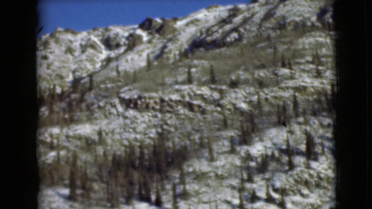 1946 Alaska 8mm frame showing a snow-patched rocky mountainside with scattered dark trees and a clear blue sky.