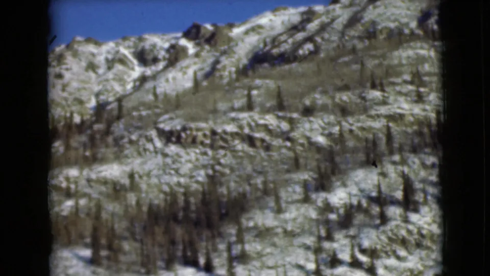1946 Alaska 8mm frame showing a snow-patched rocky mountainside with scattered dark trees and a clear blue sky.
