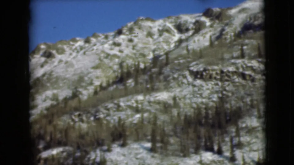 1946 Alaska home-movie frame of a rugged snow-dusted mountain slope, sparse trees, and open blue sky.