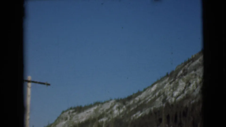 1946 Alaska 8mm frame showing a wooden telephone pole near evergreen slopes and snow-streaked mountains under clear sky.
