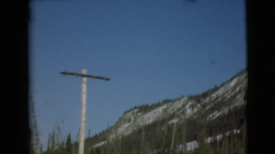 1946 Alaska home-movie frame with a wooden utility pole centered before a rocky, snow-dusted mountain ridge.