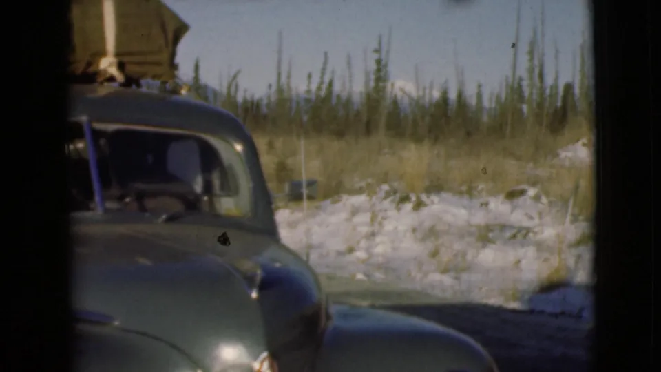 1946 Alaska frame with a dark rounded car approaching the frame, its roof loaded with canvas-wrapped gear near snowy roadside trees.
