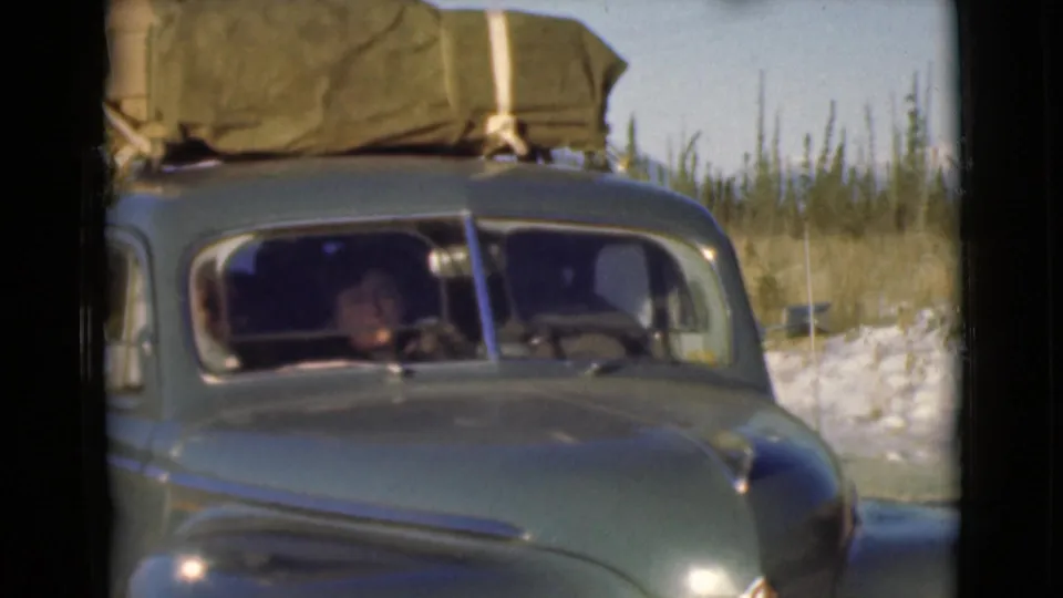 1946 Alaska home-movie frame showing a loaded dark car at close range, with a seated woman visible inside through the windshield.