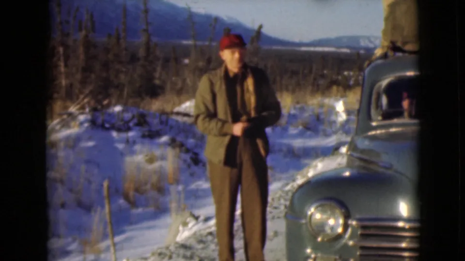 1946 Alaska frame showing a man in winter clothing beside a dark packed car, with snow, trees, and mountains in the background.