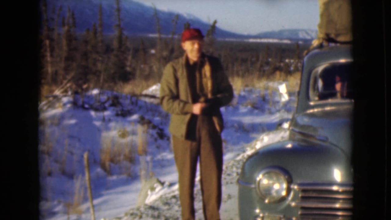 In supported 1946 Alaska footage, a man stands near a green vintage car with snow, trees, and mountains behind him.