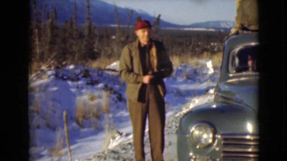 A man in winter clothing stands by a green vintage car in a snowy Alaska landscape, with trees and mountains visible.