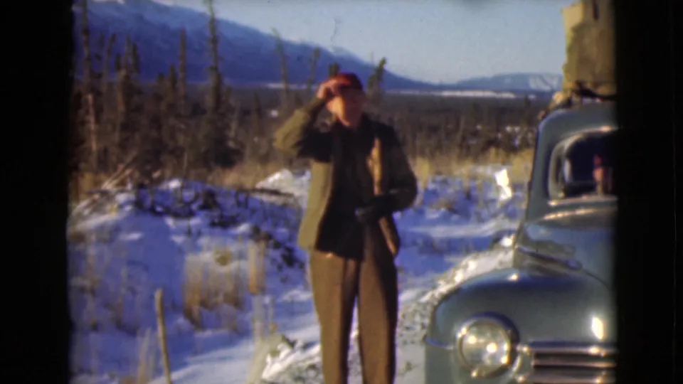 The man appears to adjust his red cap beside the green car, with snowy ground and distant mountains in view.