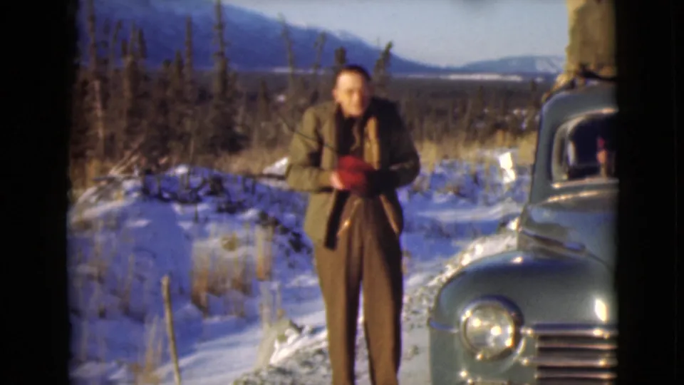 A man beside a green vintage car handles his red cap or clothing while standing in a snow-covered Alaska scene.