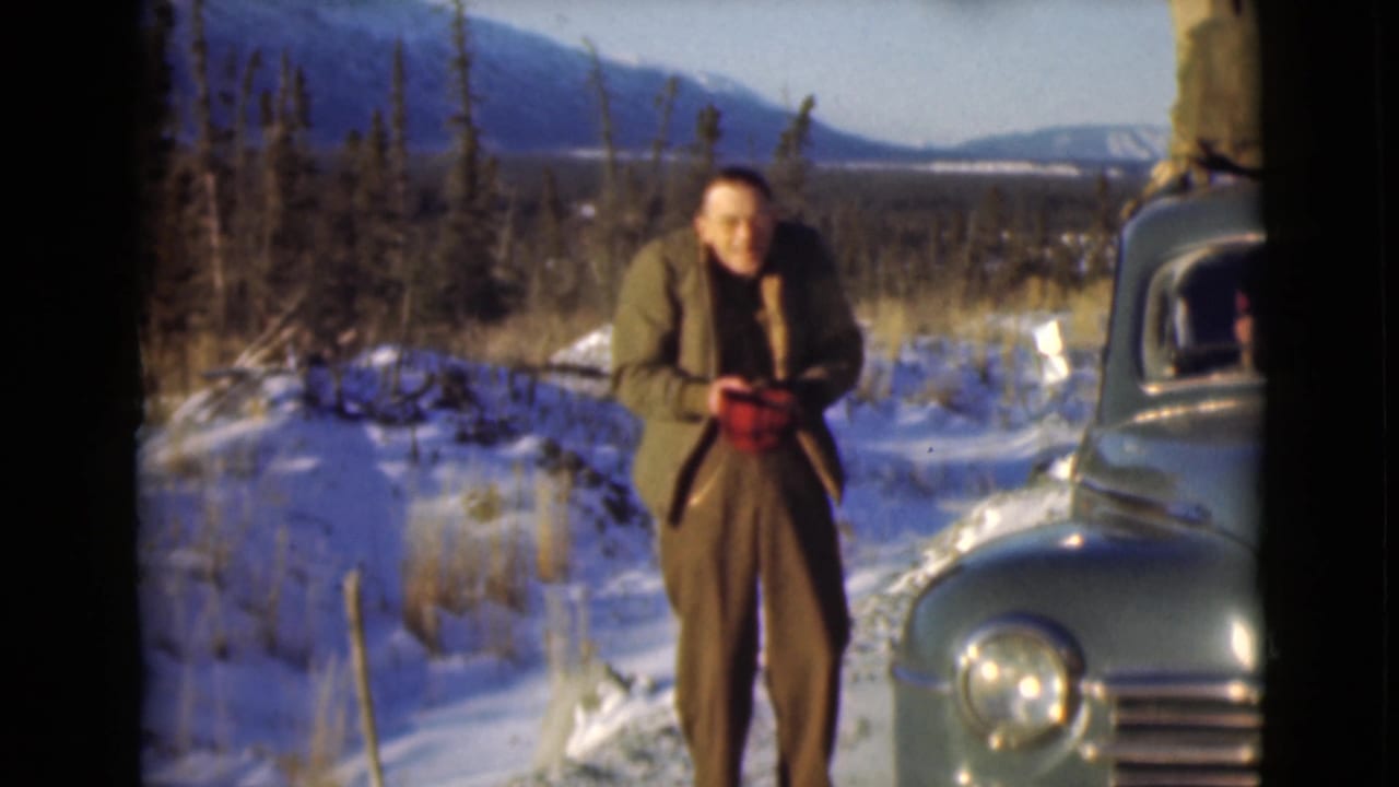 Man beside a blue vintage car on a snowy roadside with mountains in the distance.