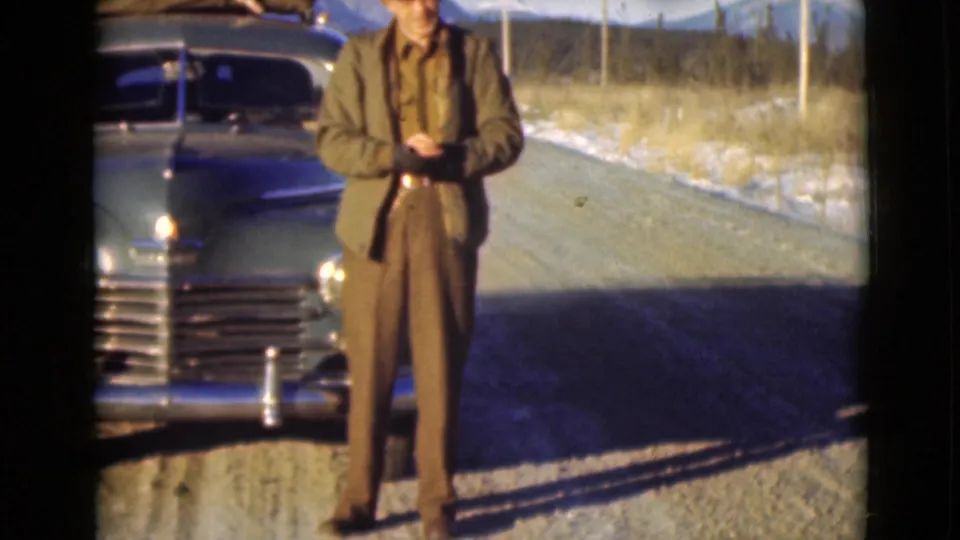 A man in brown jacket and trousers stands before a blue vintage automobile on a snowy Alaska roadside, holding a red cap.