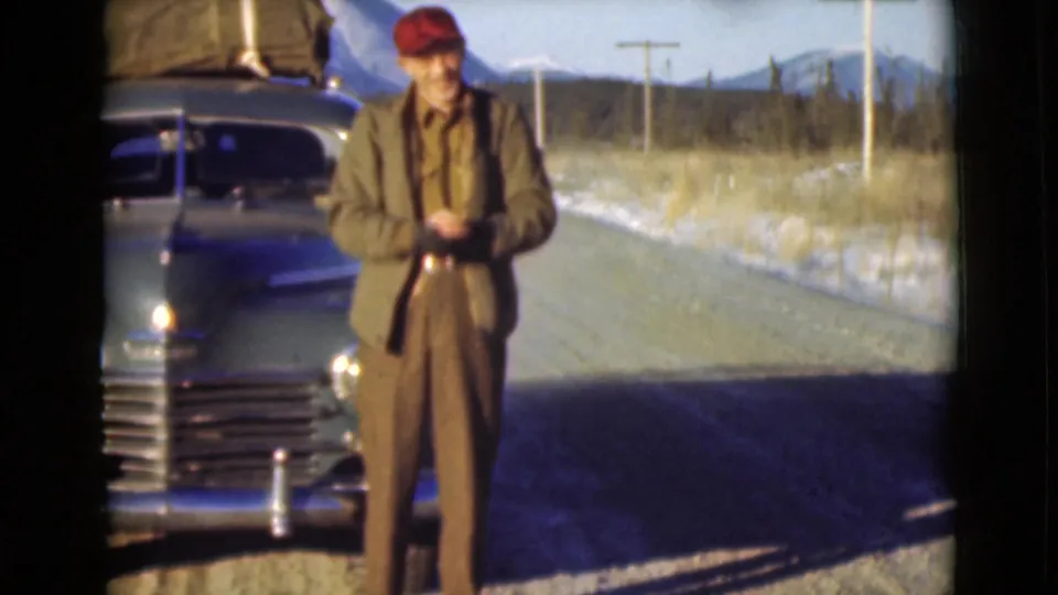 The man, now wearing the red cap, stands near the grille of a blue vintage car on a cold roadside in Alaska.