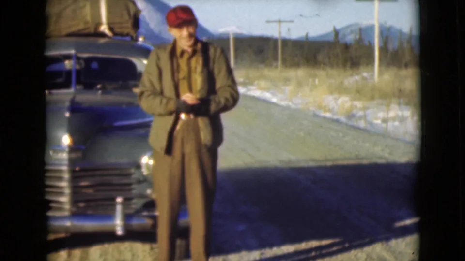 A posed home-movie frame shows the man in a red cap beside the blue car, with snow, road, trees, and mountains in the background.